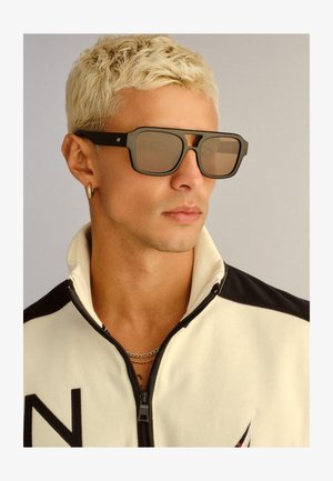 Blonder Mann mit kurzen Haaren, der große schwarze Sonnenbrillen, kleinen goldenen Creolen, mehrere Ketten und eine weiße Jacke mit Reißverschluss und schwarzen Akzenten trägt.