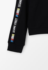 Manica di felpa nera con polsino a coste. Presenta strisce verticali con "PUMA" in blu e "SESAME STREET" in bianco, oltre a dettagli multicolore.