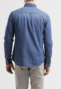 Camisa de mezclilla en azul medio, de manga larga con cuello abotonado, que presenta una sutil textura y un dobladillo curvado. La vista trasera muestra los detalles de costura.