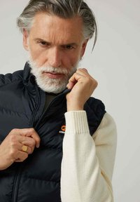 Chaleco negro acolchado sobre un suéter de punto crema; cuenta con un cuello alto y un sutil logo de marca en el lado izquierdo del pecho; textura suave.