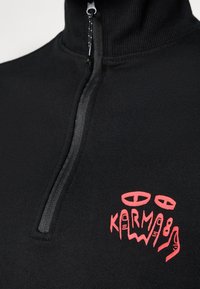 Black zip-up sweater made of stretch fabric, featuring a high collar and prominent red graphic text on the chest. Smooth texture, fitted design.