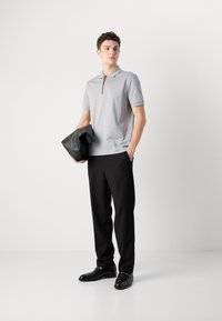 Sivá polo košeľa s krátkymi rukávmi a zipsovým golierom, skombinovaná s čiernymi nohavicami a čiernymi koženými topánkami. Model drží čiernu tašku.