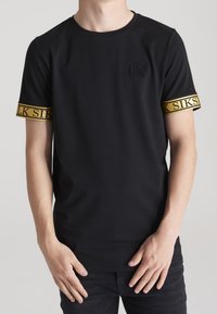 La camiseta negra de manga corta cuenta con un cuello redondo, un logo bordado y acentos dorados en las mangas con texto. Tejido suave y liso.