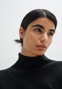 Prim-plan al unei persoane care poartă un pulover negru cu guler înalt. Prezintă o piele netedă, păr închis la culoare și sprâncene bine definite pe un fundal deschis.