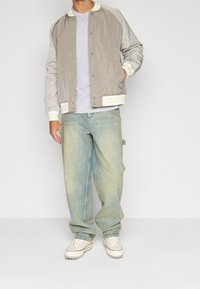 Veste bomber légère en beige avec des manches gris clair, poignets rayés ; associée à un jean baggy bleu délavé et des baskets blanches.