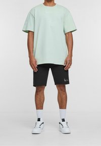Jasnozielona, oversize'owa koszulka z krótkim rękawem i białym logo umieszczonym na środku klatki piersiowej, w zestawie z czarnymi szortami i białymi trampkami.