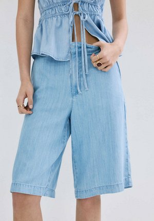 Donna che indossa pantaloncini bermuda in denim azzurro chiaro e una canotta senza maniche abbinata con coulisse in vita, mostrando anelli sulle dita.