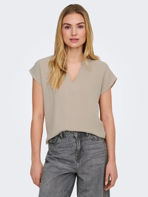 Vrouw met een beige top met korte mouwen en V-hals, en hooggetaillede vervaagde zwarte spijkerbroek, staand tegen een lichtgrijze achtergrond.