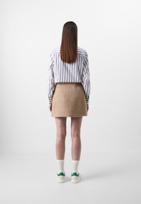Gestreepte blouse met lange mouwen, beige wollen rok, witte sokken, schoenen met groene accenten; bekeken van achteren tegen een effen achtergrond.