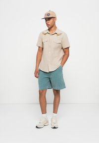 Beige short-sleeve shirt with embroidered accents, paired with teal denim shorts. Wearing a beige cap and chunky beige sneakers with white socks.