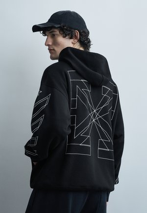 Νέος άνδρας με μαύρο καπέλο και φούτερ με κουκούλα, με λευκό γεωμετρικό σχέδιο σε γραμμή στην πλάτη και τα μανίκια, στέκεται μπροστά σε απλό φωτεινό γκρι φόντο.