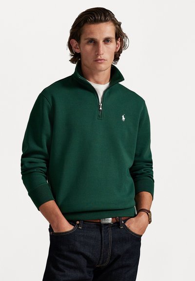 Groene rits-sweater met hoge kraag en geribde manchetten, in combinatie met donkere spijkerbroek. Opvallend wit logo op de borst.