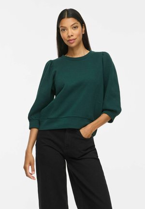 Donna con lunghi capelli scuri che indossa una maglietta verde scuro con maniche a sbuffo e pantaloni neri, in piedi con una mano in tasca su sfondo bianco.