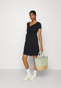 Vestido preto de manga curta em estilo envoltório com decote em V, acompanhado de uma bolsa de palha tecida em verde e natural. O modelo usa meias brancas e ténis.