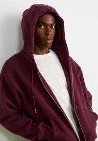 Bordeaux lynlås hættetrøje med stor hætte, blød tekstur og frontlommer, lagt over en lysecreme t-shirt med et diskret logo-print.