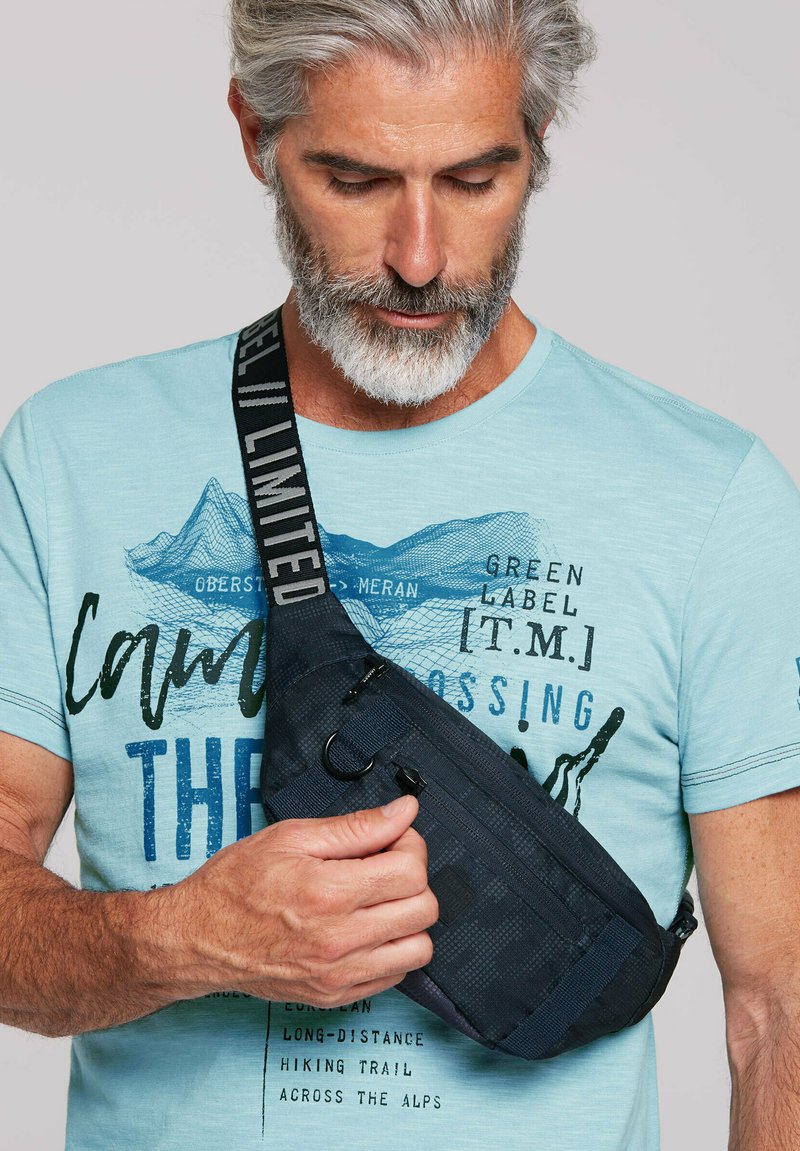 Schwarze Umhängetasche aus strukturiertem Stoff, mit verstellbarem Gurt mit dem Text "LIMITED" und Reißverschluss, quer über der Brust getragen.