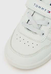 Baskets blanches en cuir avec un bout perforé, des lacets et une sangle Velcro. Logo "TOMMY" en bleu foncé au-dessus de la sangle. Surface texturée et accents cousus.