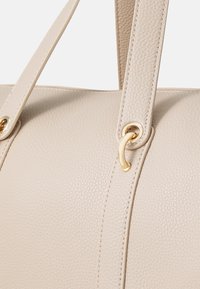 Beige leather handbag with a textured finish, featuring long straps and gold-toned hardware accents near the attachment points.
