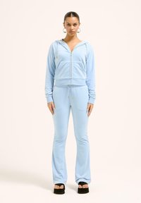 Šviesiai mėlyna zip-up kapučino su priekinėmis kišenėmis ir raišteliais, suderinta su atitinkančiomis sportinėmis kelnėmis. Medžiaga atrodo minkšta su lygia tekstūra.