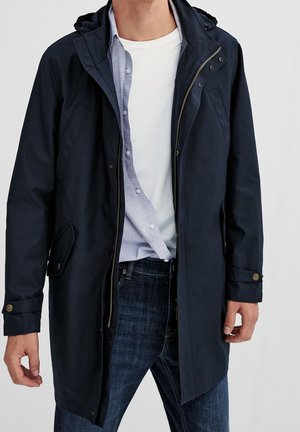 Mand iført navy hættetrøje over lyseblå skjorte med knapper og hvid t-shirt, kombineret med mørkeblå jeans.