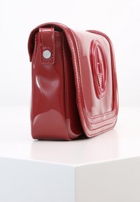 Borsa a tracolla in pelle rossa lucida con logo impresso sul pattina, dettagli in bottone argentato sul lato, esposta su una superficie bianca.