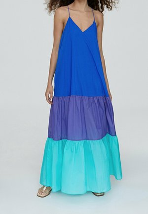 Femme portant une robe longue sans manches à trois niveaux avec des blocs de couleur bleu, violet et turquoise et fines bretelles, debout pieds nus.