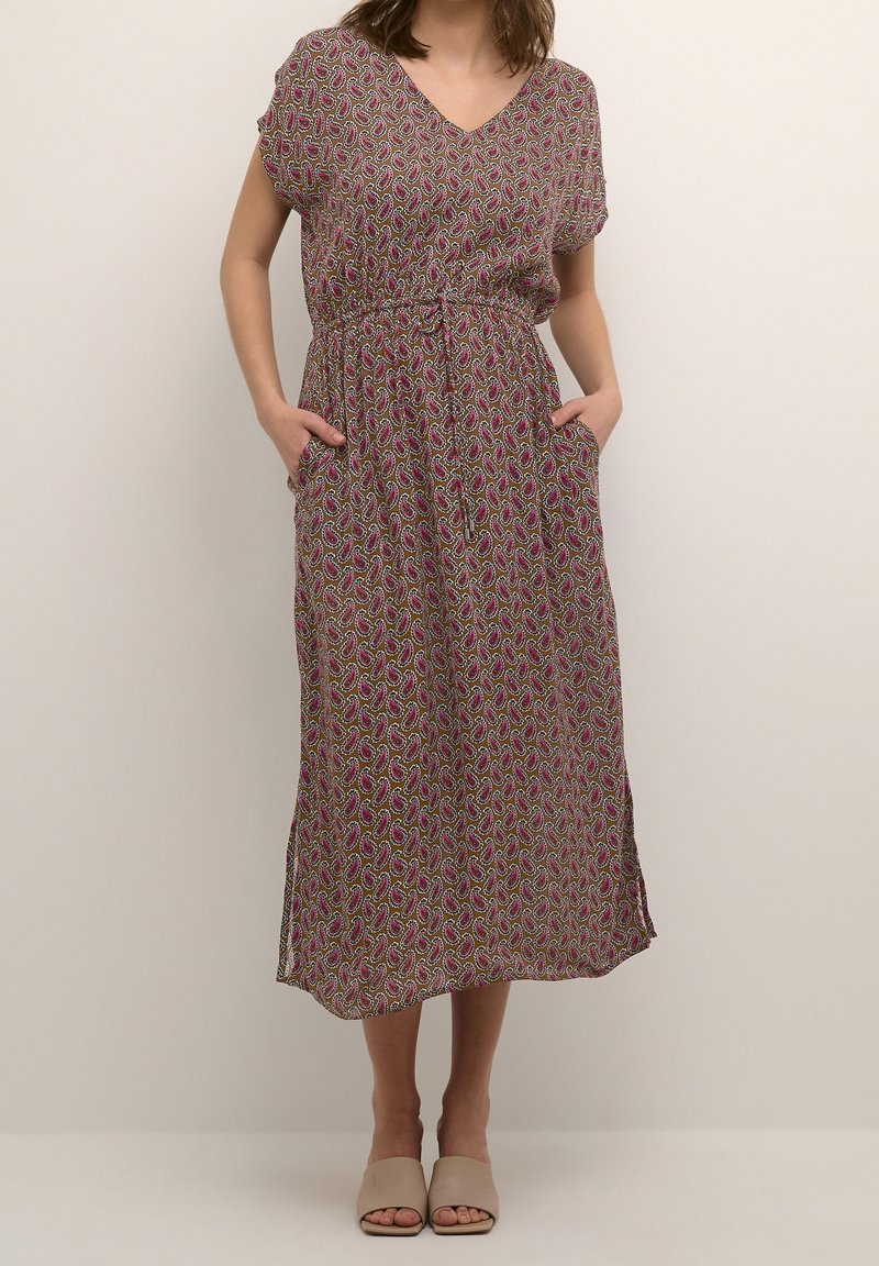 Femme portant une robe midi à motif cachemire rose et manches courtes, les mains dans les poches, associée à des mules ouvertes beiges.