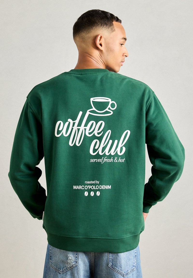 Groene sweatshirt met witte tekst die luidt "koffieclub vers en heet geserveerd," met een koffiekop-illustratie en merknaam eronder.