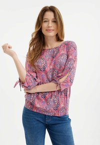 Rosa Bluse mit Paisley-Muster und Dreiviertelärmel, mit Bindebändern und lockerer Passform. Kombiniert mit blauen Jeans.
