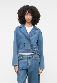 Izgriezta zila džinsu jaka ar divrindu dizainu, lieliem uzklājiem un jostas detaļām. Valkāta virs baltas krekla un kombinēta ar džinsiem.