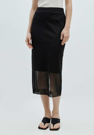 Jupe midi noire avec une taille ajustée, une couche inférieure transparente et une silhouette droite. Associée à des tongs noires.