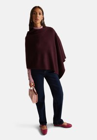Donna che indossa un poncho viola scuro, un dolcevita rosa chiaro, jeans blu scuro e ballerine rosa, mentre tiene una piccola borsa rosa ed è in piedi su uno sfondo bianco.