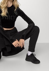 Černý sametový top s dlouhým rukávem a zkrácenou délkou se textem "Juicy", spárovaný s černými tepláky s malým fialovým logem. Černé tenisky dotvářejí celý outfit.