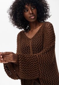 Brązowy, ażurowy sweter z dekoltem w serek i szerokimi rękawami, o luźnym, teksturowanym wzorze i widoczną skórą pod spodem.