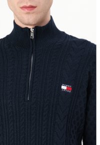 Tengerészkék kötött pulóver magas nyakkal, negyed cipzárral és texturált kábelmintákkal, a mellrészen kis 'Tommy Jeans' logóval.