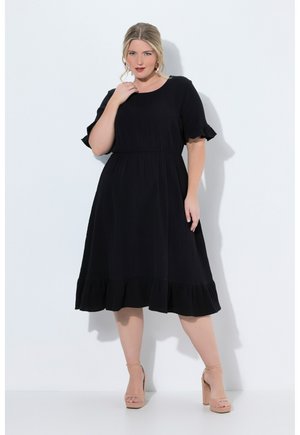 Donna taglia forte che indossa un vestito nero al ginocchio con maniche e orlo arricciati, posando davanti a uno sfondo bianco semplice.