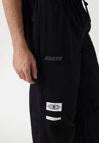 Sorte stof joggers med et "GUESS" logo patch, sidelomme og hvide accentmærkater med grafiske designelementer.