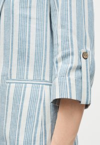 Κοντινό πλάνο ενός μανικιού με κάθετες μπλε και λευκές ρίγες, ξύλινο κουμπί και τσέπη πλαϊνή με σκίσιμο, σε ένδυμα.