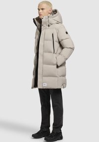 Chaqueta acolchada beige con capucha, que presenta un cierre de cremallera, dos bolsillos laterales con cremallera y acentos en negro. Llevada sobre pantalones vaqueros oscuros y zapatos negros.