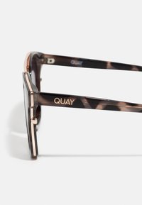 Slnečné okuliare v korytnačkovom vzore s čiernymi šošovkami. Majú kovové zlaté akcenty a charakteristický štvorcový tvar. Názov značky "QUAY" je výrazne zobrazený.