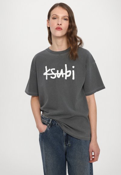 Tricou oversized gri cu un logo alb "ksubi" pe față. Dispune de mâneci scurte și un guler rotund, asortat cu blugi albaștri.