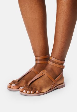 Hnědé kožené sandály s designem T-pásku, s popruhy kolem kotníků a plochou podrážkou. Vyznačují se jednoduchou, minimalistickou estetikou s matným povrchem.