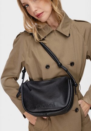 Vrouw in beige trenchcoat met een zwarte leren crossbodytas met het geprofileerde merknaam "PIKOLINOS" aan de voorkant.