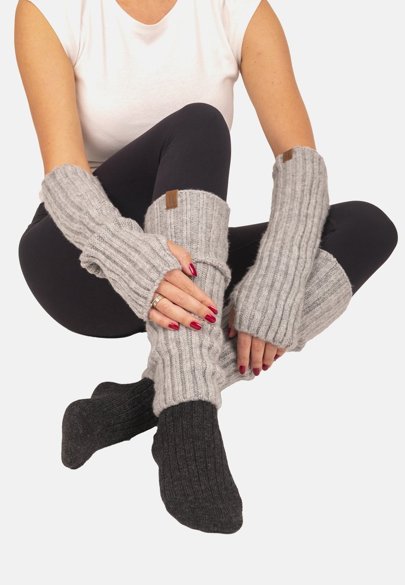 Mujer sentada con las piernas cruzadas, lleva leggings negros, camiseta sin mangas blanca, calentadores de brazo de punto gris, calentadores de piernas a juego y calcetines gris oscuro.