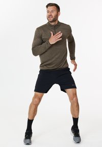 Langärmliges, olivgrünes Sportoberteil mit einem Viertelreißverschluss-Design, kombiniert mit schwarzen Shorts. Graue Laufschuhe und schwarze Kniesocken.