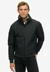 Chaqueta bomber negra con cremallera frontal y cuello alto, que presenta un acabado texturizado y puños acanalados. Dos bolsillos laterales visibles.