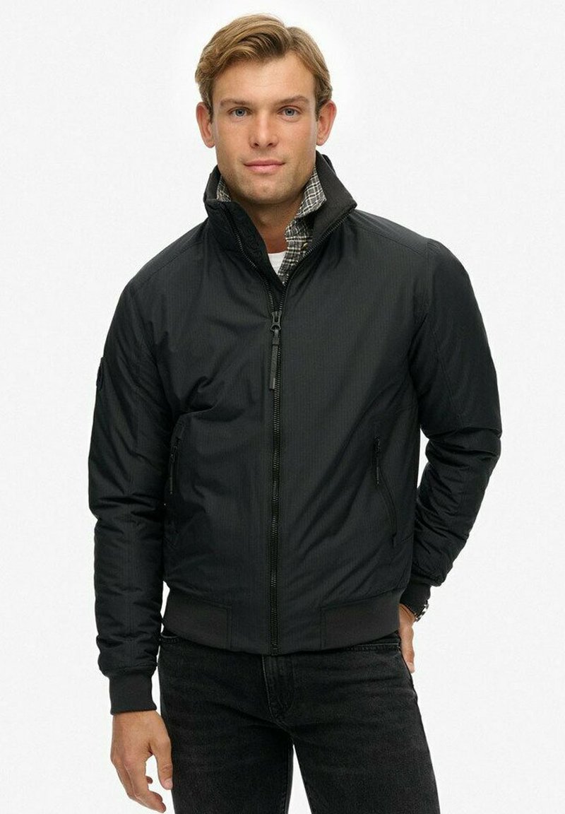 Chaqueta bomber negra con cremallera frontal y cuello alto, que presenta un acabado texturizado y puños acanalados. Dos bolsillos laterales visibles.