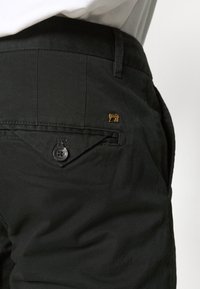 Pantalons en tissu noir avec une poche arrière dotée d'une fermeture à bouton et d'un petit logo doré situé au-dessus de la poche. Texture lisse.