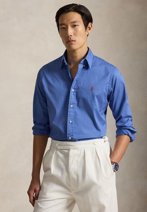 Homme portant une chemise bleue boutonnée avec les manches retroussées et un pantalon blanc plissé, debout avec une main dans la poche sur un fond neutre.