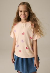 Helles Pfirsichfarbenes Baumwoll-T-Shirt mit kurzen Ärmeln, das auf der Vorderseite mit gestickten Schmetterlingen in rosa, lila und gelben Mustern versehen ist.